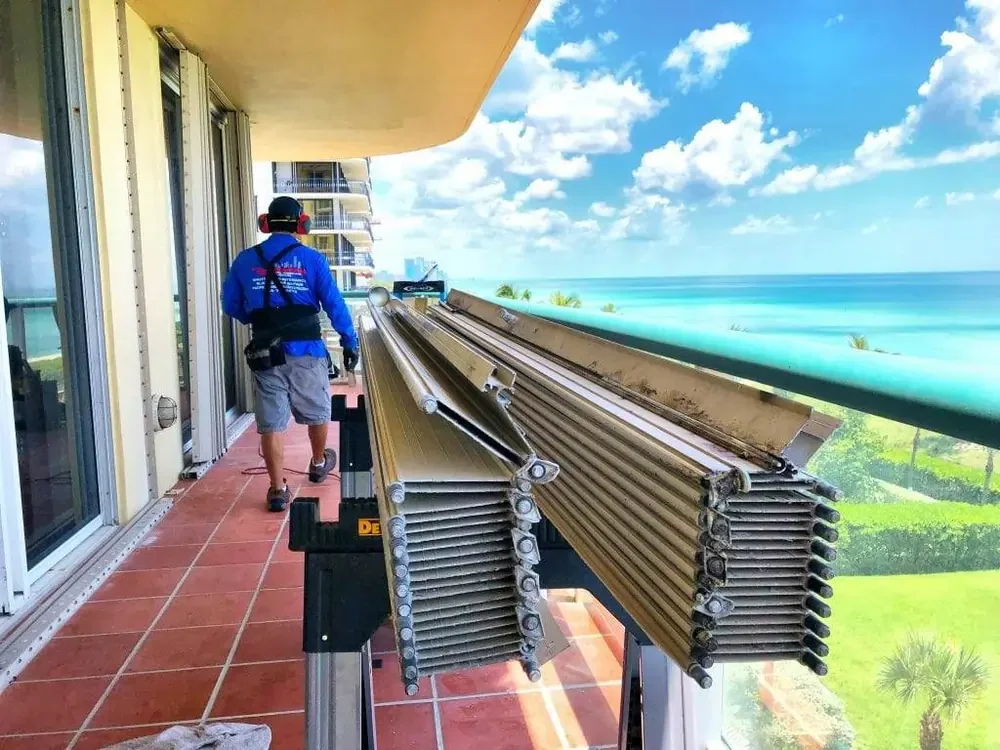 Worker installing accordion hurricane shutters on coastal high-rise balcony with ocean view