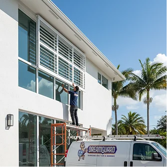 Window technician on ladder cleans glass façade beside branded service van in tropical setting.